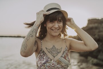 Canvas Print - Young female with tattoos wearing a dress and straw hat on blurred ocean background