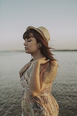 Wall Mural - Young female with tattoos wearing a dress and straw hat on blurred ocean background