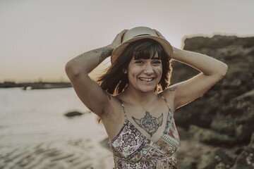 Wall Mural - Young female with tattoos wearing a dress and straw hat on blurred ocean background