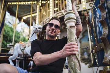 Canvas Print - Young Caucasian male spending fun time in an attraction park with a carousel with horses at daytime