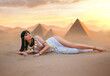 Egypt Style Rich Luxury Woman. Sexy beautiful girl goddess Queen Cleopatra lies on yellow sand desert pyramids. Art ancient pharaoh costume white dress gold accessories Black hair wig Egyptian makeup