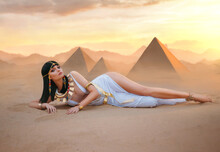 Egypt Style Rich Luxury Woman. Sexy Beautiful Girl Goddess Queen Cleopatra Lies On Yellow Sand Desert Pyramids. Art Ancient Pharaoh Costume White Dress Gold Accessories Black Hair Wig Egyptian Makeup