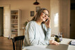 © shurkin_son - Positive cheerful mature woman pensioner in rectangular eyeglasses using laptop pc sitting at table in cozy home interior, smiling, speaking to friend on smart phone, discussing latest news