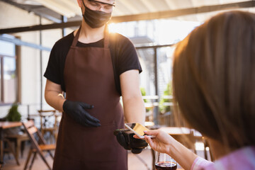 Wall Mural - The waiter works in a restaurant in a medical mask, gloves during coronavirus pandemic. Representing new normal of service and safety. Gets paid order with device contactless. Blank screen for your ad