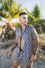 Poster - Male with tattoos on his chest, arms, and legs wearing fashionable clothes posing at the beach