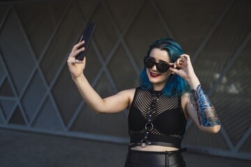 Canvas Print - Europian female with tattoo and blue hair wearing sunglasses and sexy black dress taking a selfie