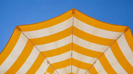 White and yellow striped beach umbrella. Blue sky in the background. View from below. Relaxing context. Summer holidays by the sea. General contest and location