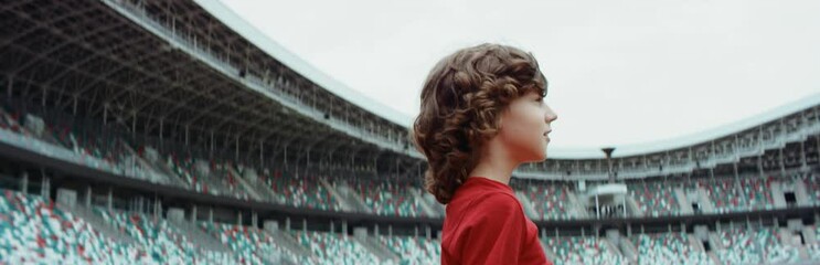 Wall Mural - Cute little kid boy soccer player standing on an empty stadium, dreaming of becoming professional player, soccer star. RED cinema camera RAW graded footage