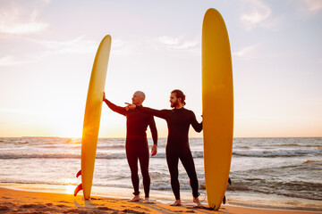 Two young surfers in black wetsuit with yellow surfing longboards on a ocean coast at sunset ocean. Water sport adventure camp and extreme swim on summer vacation.