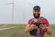 Young worker with strong beard in wind park. Holding tablet. Wind Mill  in background.