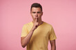 © timtimphoto - Severe young handsome short haired brunette man raising hand with hush gesture to his mouth and frowning eyebrows while looking seriously at camera, isolated over pink background