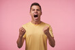© timtimphoto - Expressive young handsome brown haired male raising excitedly his fists and shouting emotionally with wide mouth opened, isolated over pink background