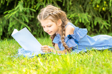 a little girl with pigtails reads a book lying on the green grass in the park. copy space.