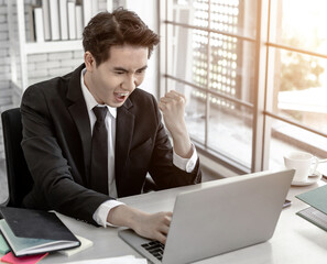 Happy of asian young businessman see a successful business plan on the laptop computer and pen on wooden table background in office,business expressed confidence embolden