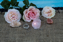 On Jute Fabric Are Semiprecious Stones Rose Quartz, Dolomite, Halite. Nearby Are Silver Rings And A Chain. In The Background There Are Three Pink Roses..