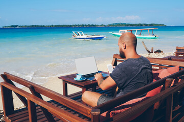 Fototapete - Businessman in casual wear working online during vacation  using netbook and wireless internet, male millennial holding mobile phone while checking booking reservation in app sitting on beach .