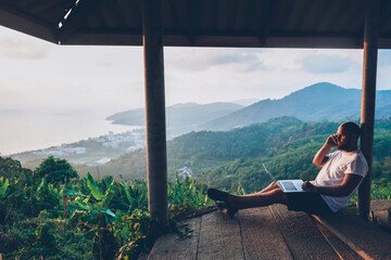 Hipster gut traveler working remote while enjoying Thailand nature landscape during summer vacations. Male freelancer using internet connection and modern devices outdoors against tropical environment