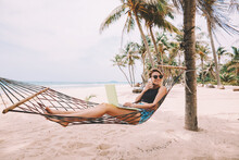 Successful Female Tourist In Sunglasses Talking On Smartphone In Roaming During Vacations While Work Remotely At Modern Laptop Computer Lying On Hammock On Sea Shore Of Tropical Island With Palm Trees
