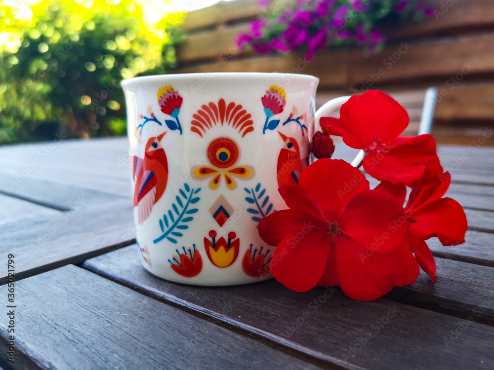 Piękny folklorystyczny kubek do kawy ręcznie malowany postawiony na drewnianym blacie w otoczeniu kwiatów pelargonii. - obrazy, fototapety, plakaty 