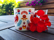 Piękny folklorystyczny kubek do kawy ręcznie malowany postawiony na drewnianym blacie w otoczeniu kwiatów pelargonii.
