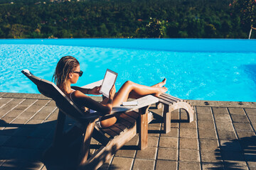 Wall Mural - Sexy young woman sunbathing on sunbed near blue pool while reading news on websites using good 4G internet connected to digital tablet with blank screen area during summer tropical resort