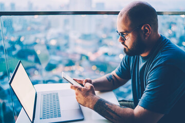 Fototapete - Young handsome skilled IT professional in cool spectacles chatting with friends in social network via smartphone.Good looking bearded male entrepreneur texting email message on modern cellular