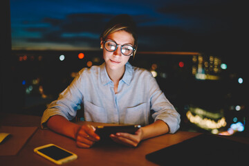 Wall Mural - Young woman staying late watching webinar on modern digital tablet sitting at desktop at home, hipster girl reading book online on portable pc sitting at home interior with window night cityscape