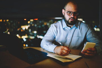 Fototapete - Concentrated businessman noting contact information from cellphone to notepad working overtime,pensive male freelancer checking mail on smartphone while creating publication for blog in notepad