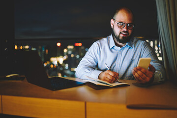 Fototapete - Cheerful businessman happy reading good news from smartphone staying lata with work, prosperous male blogger excited with good feedbacks on web site on cellular creating new publication in netbook