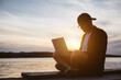 © mykolastock - Silhouette of modern young hipster male entrepreneur wearing baseball cap and eyeglasses working on laptop near lake at sunset