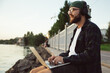 © mykolastock - Modern handsome cheerful young bearded hipster freelance developer man siiting and working on laptop on river bank.
