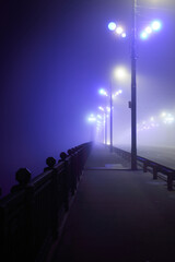 Wall Mural - Pedestrian walkway through the illuminated empty Stone bridge in a thick fog at night. Lanterns close-up. Daugava river, Riga, Latvia. Concept image, neon colors