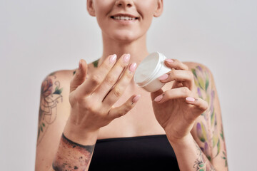 Cropped portrait of smiling tattooed woman trying skin care product, holding white plastic jar of cream or body lotion isolated over grey background