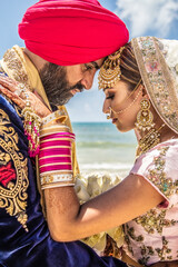 Wall Mural - Sikh bride and groom wearing bright traditional clothing on clear sandy beach beneath sunny blue skies