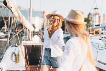 Two beautiful blonde girls friends mother and daughter in white and straw hats on the yacht at the pier