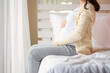 © ueaphan - Young beautiful pregnant woman sitting on bed and touching belly near the window in bedroom. Pregnancy, motherhood, people and expectation concept.
