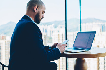 Fototapete - Pensive bearded director in elegant suit sending notification with important information on smartphone device sitting at lapto computer wth mock up area for advertising text in office interior