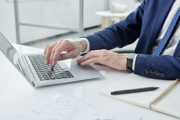 Poster - Close-up of unrecognizable businessman in jacket sitting at desk with notepad and typing on laptop in office