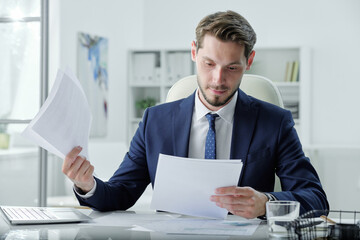 Wall Mural - Busy young manager in formal suit sitting at desk and examining business papers in office