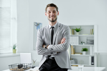 Poster - Portrait of smiling confident young businessman in black tie standing with crossed arms near desk in office