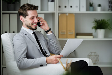 Wall Mural - Cheerful successful young businessman with beard sitting in comfortable armchair in office and viewing contract while communicating by phone