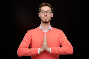 Wall Mural - Serene young man with stubble holding hands in Namaste while meditating with closed eyes against black background