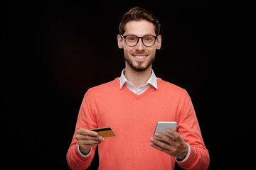 Poster - Portrait of cheerful handsome young man with stubble standing with credit card and smartphone and using online transaction service