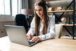 © Kateryna - A beautiful caucasian female student is studying in college remotely. She is sitting at a table at home with a laptop and concentrated is watching a video conference lesson