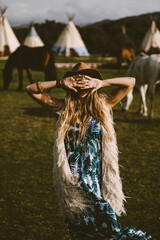 A blonde bohemian woman with a hat outdoors in a meadow with horses background