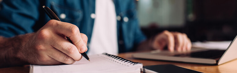 panoramic crop of businessman writing in notebook near smartphone and laptop