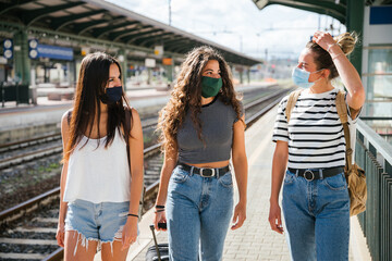 Three young friends women at the station walking and waiting train for their trip in summer with face mask for protection by infection from Coronavirus, Covid-19 - Millennials having fun together