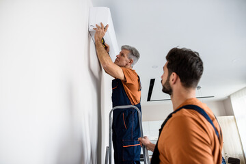 Two workers in uniform, air conditioning masters using ladder while installing a new air conditioner in the apartment. Construction, maintenance and repair concept