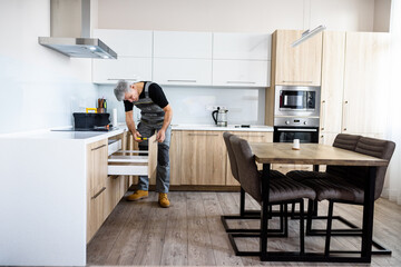 Full length shot of aged repairman in uniform working, fixing kitchen cabinet using screwdriver. Repair service concept