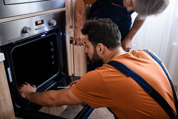 Close up shot of two repairmen, workers in uniform examining oven using screwdriver and flashlight indoors. Repair service concept